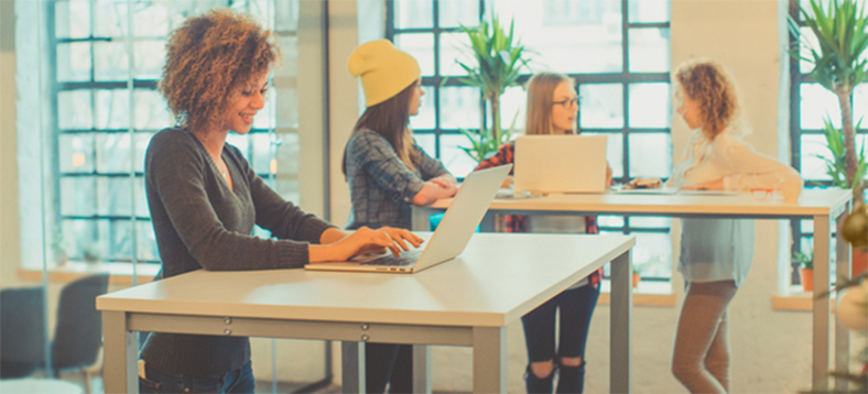 Four young women using standing desks. One is working alone on one desk with a laptop