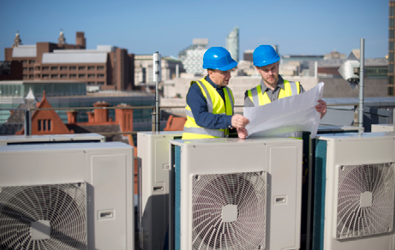 two men with safety helmets and vests on rooftop looking at a document surrounded by system fans