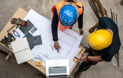 overhead view of two people wearing construction helmets pointing to a project document along with other tools spread out on a table