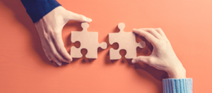 one hand holding a puzzle piece trying to connect with another puzzle piece held by another hand