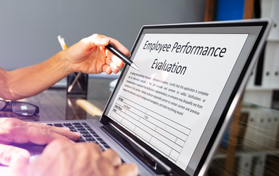 hand with pen pointing to a laptop screen with an Employee Performance Evaluation document open while another set of hands are typing on the laptop