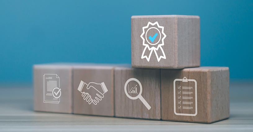 Wooden blocks with icons of checklist, handshake, analytics, and certification badge, symbolizing business quality assurance, partnership, performance evaluation, success strategy, and corporate achievement.