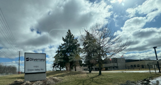Photograph of Diversey facility exterior showing the Diversey street sign and address "1151 Green Valley Rd." in front of the building under a partly cloudy sky. The scene includes leafless and evergreen trees, a grassy area with patches of snow, and utility poles along a road.