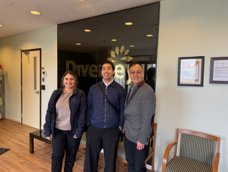 Photograph of three OECM Staff inside the Diversey facility front entrance. The setting includes wooden flooring, a bench, and two framed documents on a light-colored wall with backdrop of Diversey logo.