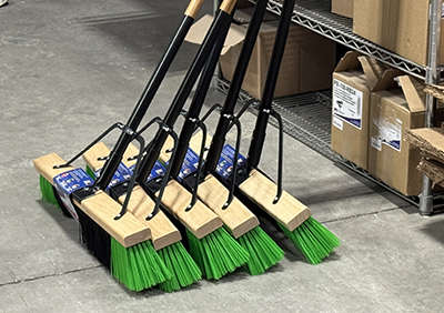 Row of industrial push brooms with green bristles lined up on a warehouse floor