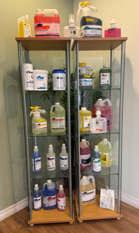 Photograph of two glass display cabinets filled with various cleaning and disinfectant products, including spray bottles, jugs, and containers in different colors such as yellow, pink, blue, and white. Labels on products indicate brand names and usage instructions, with cabinets positioned against a light-colored wall and wooden floor.